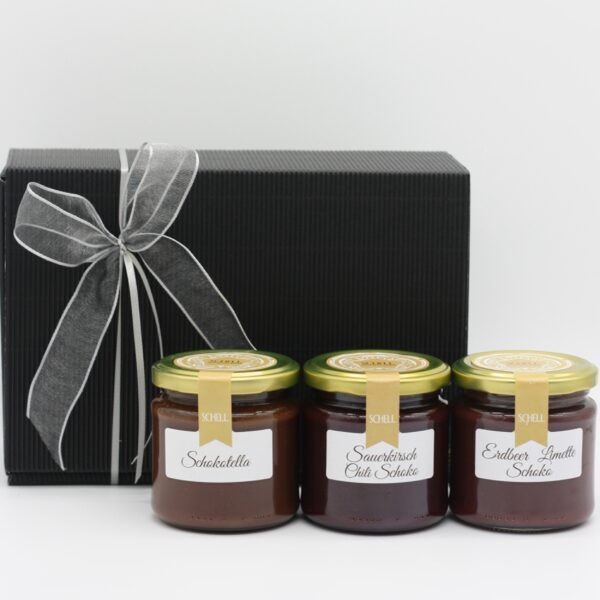 Schokofrühstück Karton als Geschenk verpackt im Hintergrund. Im Vordergrund sehen wir ein Glas Schokotella, ein Glas Sauerkirsch Chilli Schoko und ein Glas Erdbeer Limette Schoko.