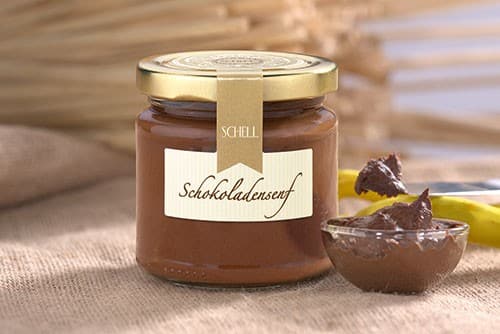 Der Schokoladensenf von Schell Schokoladen ist in einem kleinem Glas mit goldenem Deckel, Verschlusssicherung und einem Etikett , daneben steht eine kleine Glasschale mit Schokosenf gefüllt