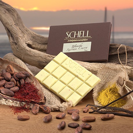 Die verpackte Schokolade lehnt an ein geschwungenes Holz, darunter die nicht verpackte weiße Schokolade und eine Kakaoschote gefüllt mit Kakaobohnen und Safran. Rechts von der Schokolade befinden sich Curry und Vanilleschoten.