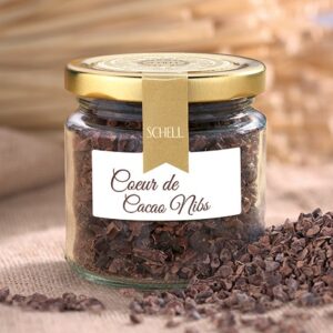 Coeur de Cacao Nibs sind in einem kleinen Glas mit goldenem Deckel, Verschlusssicherung und einem Etikett, daneben liegt etwas von den Cacao Nibs gehäuft.