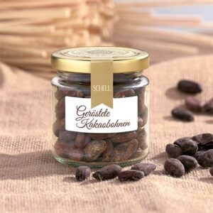 Die Geröstete Bio Kakaobohnen von Schell Schokoladen sind in einem kleinen Glas mit goldenem Deckel, Verschlusssicherung und einem Etikett, daneben liegen geröstete Kakaobohnen
