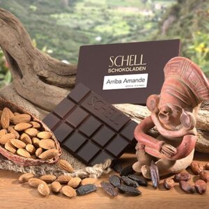 Arriba Amande in der Verpackung lehnt an ein geschwungenes Holz, darunter eine unverpackte Schokolade und eine mit Nüssen gefüllte Kakaoschote, rechts steht eine Affenfigur.