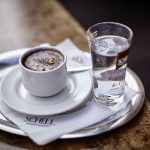 Es ist der Espresso abgebildet in einer weißen Schell Tasse und dem passenden weißen unter Teller. Zudem daneben ein Glas Wasser.