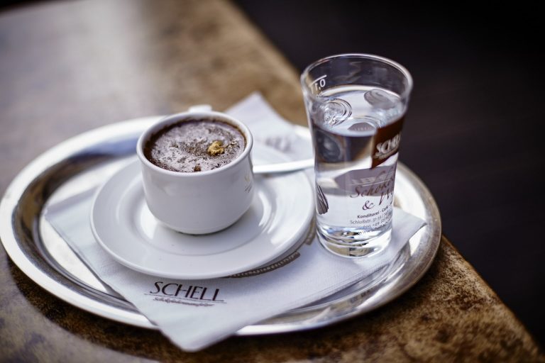 Es ist der Espresso abgebildet in einer weißen Schell Tasse und dem passenden weißen unter Teller. Zudem daneben ein Glas Wasser.