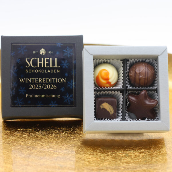 Schell Schokoladen Winteredition 2025/2026 – schwarze Pralinenschachtel und offene Vierer-Pralinenbox auf goldenem Untergrund, mit handgefertigten Pralinen in verschiedenen Formen und Dekors.