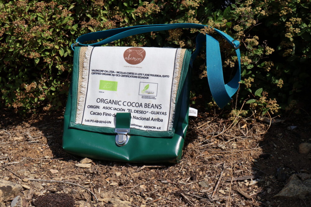 Grüne Umhängetasche Organic Cocoa Beans, die auf dem Boden steht. Im Hintergrund ist ein Gebüsch zu sehen.